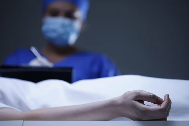 Personal médico con mascarilla tomando notas junto a un cuerpo cubierto con una sábana
