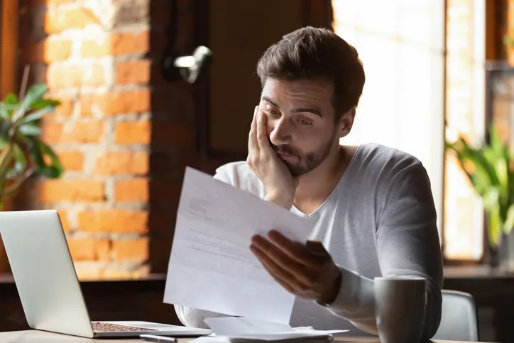 Hombre con gesto de preocupación leyendo un documento frente a su portátil, aparentando recibir una mala noticia.