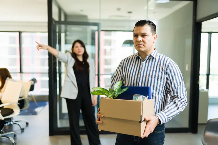 Hombre saliendo de una oficina con una caja de pertenencias mientras una mujer señala hacia la puerta, representando un despido laboral.