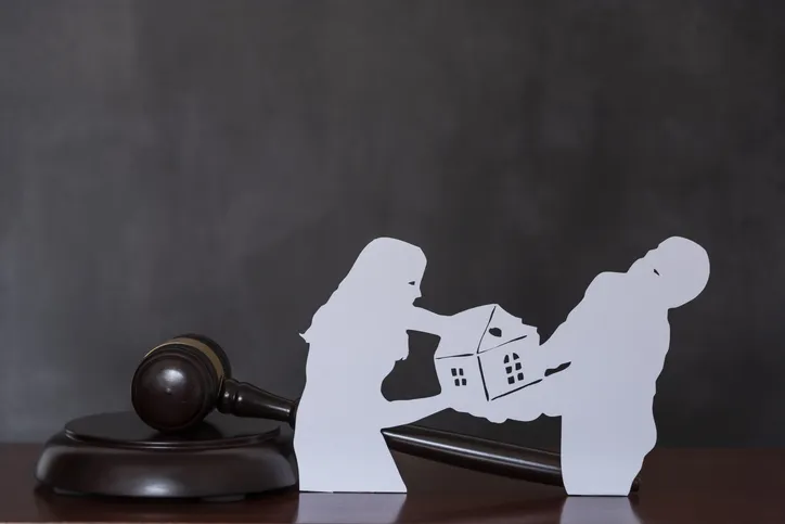 Siluetas de una pareja disputándose una casa de papel junto a un mazo de juez, representando el reparto de bienes en un divorcio