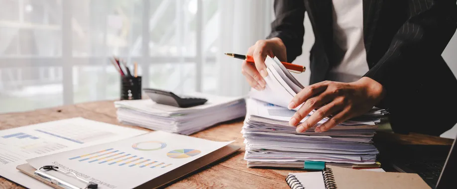 Persona revisando una pila de documentos y gráficos sobre un escritorio de oficina, junto a una calculadora y un informe con estadísticas.