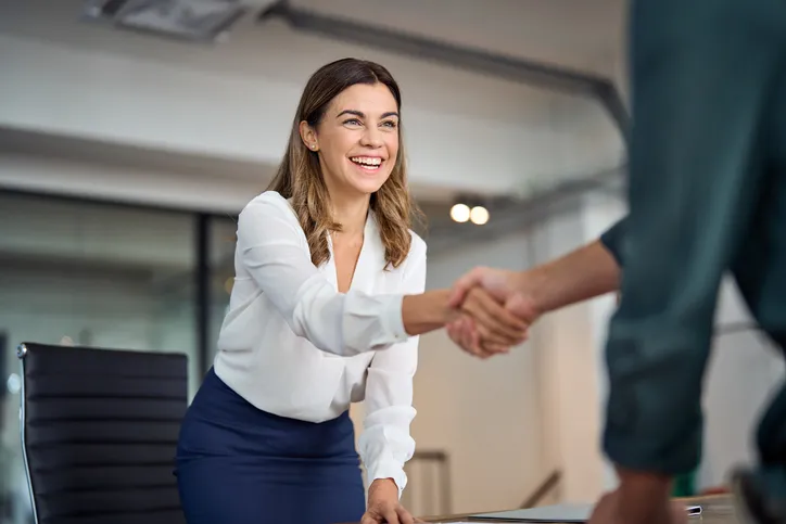 Mujer sonriente estrechando la mano de otra persona en una oficina, representando un acuerdo laboral o la contratación de un empleado.