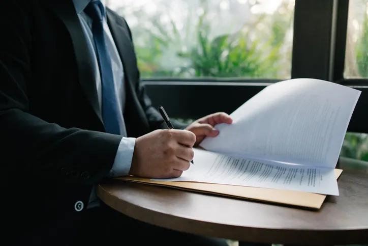 Hombre de traje revisando y firmando un documento sobre una mesa junto a una ventana con vistas a vegetación.