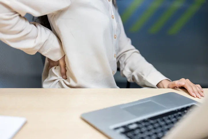 Mujer de pie junto a su portátil sujetándose la zona lumbar con la mano, mostrando dolor de espalda.