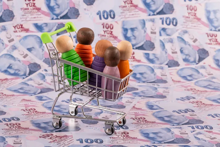 Miniaturas de personas de madera dentro de un carrito de la compra en miniatura, sobre un fondo de billetes, representando el gasto o el consumo familiar.
