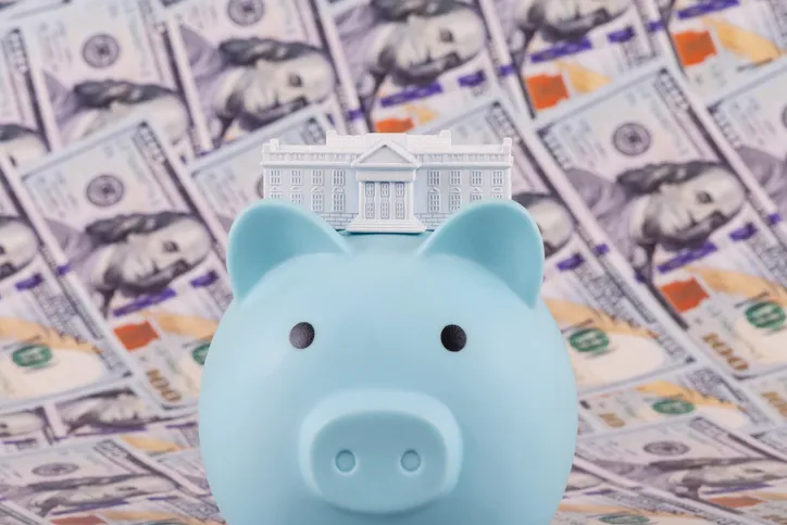 Hucha en forma de cerdito azul con una miniatura de un edificio institucional sobre la cabeza, con billetes de dólar de fondo, representando el ahorro público o las prestaciones estatales.