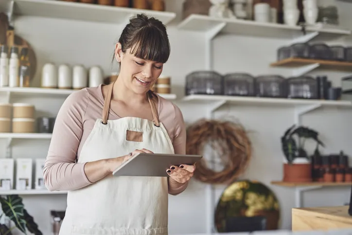Mujer con mandil sonriendo mientras usa una tablet dentro de su tienda de productos artesanales.