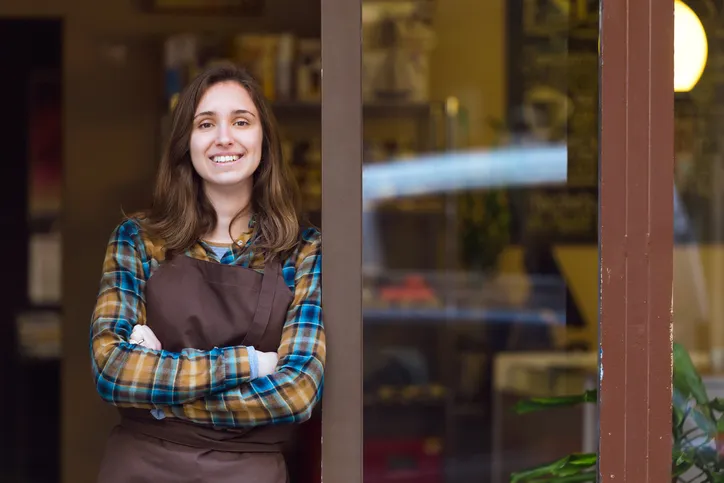 Mujer con mandil sonriendo con los brazos cruzados en la entrada de su comercio.
