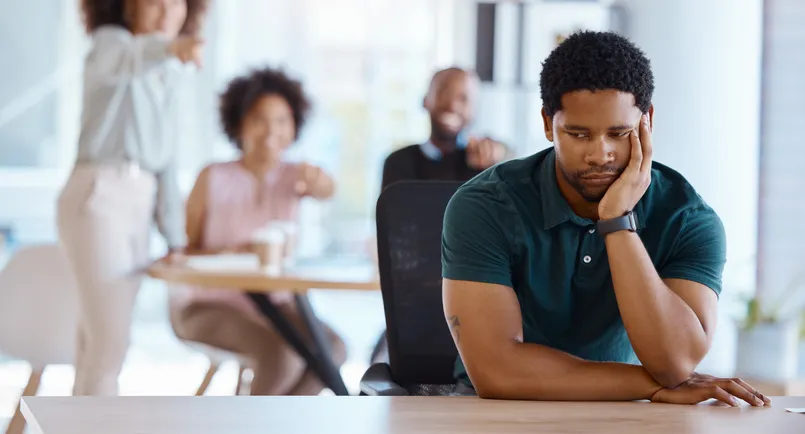 Hombre con gesto triste y aislado en una mesa mientras un grupo de compañeros se ríe y señala en el fondo, representando una situación de acoso laboral.