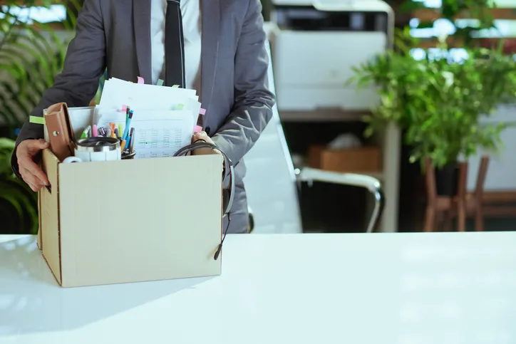 Hombre de traje sujetando una caja llena de material de oficina sobre una mesa, tras finalizar su etapa laboral.