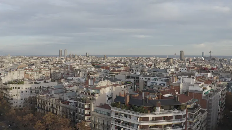 Vista aérea del skyline de Barcelona, con tejados y azoteas de edificios residenciales y el mar al fondo bajo un cielo nublado.