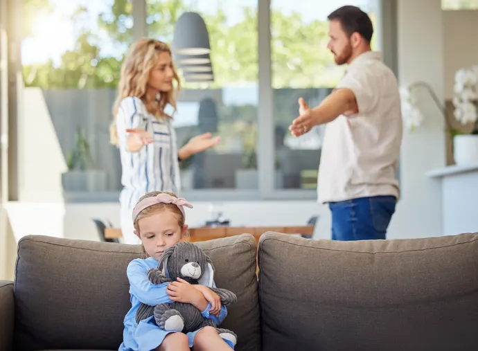 Niña triste abrazando un peluche en primer plano mientras sus padres discuten al fondo, reflejando el impacto de los conflictos en los hijos