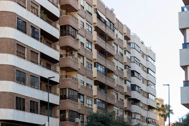 Fachada de varios edificios residenciales de ladrillo con balcones, en una calle con farolas y palmeras.