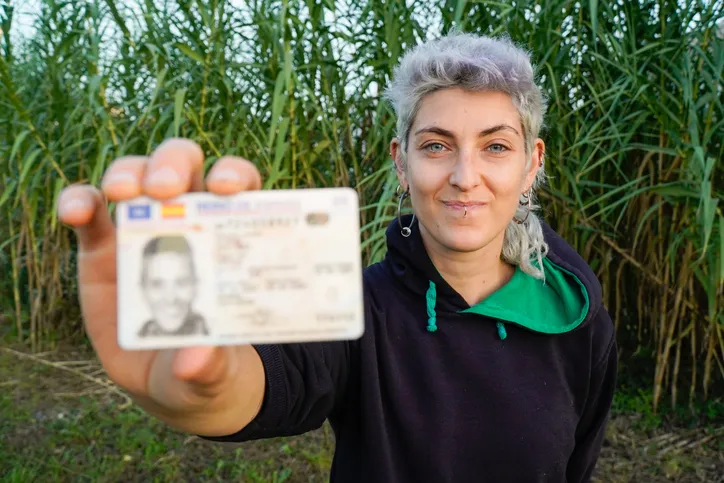 Mujer sonriente mostrando su documento de identidad español frente a la cámara, con un campo de plantas altas de fondo.