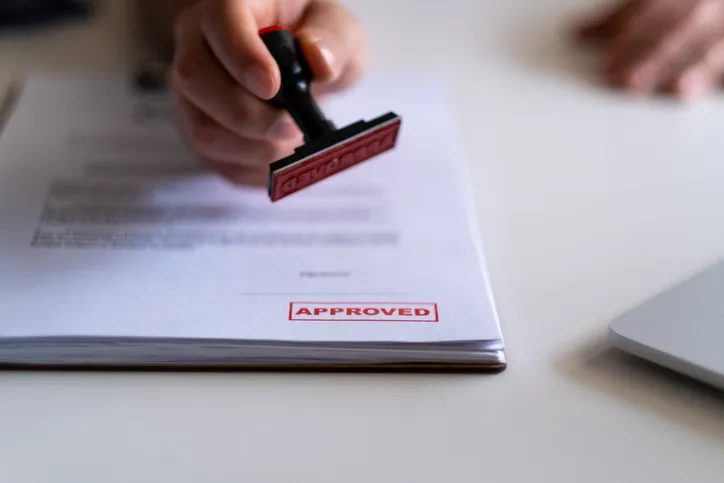Mano sellando un documento con el sello rojo 'APPROVED', indicando la aprobación de una solicitud o trámite.