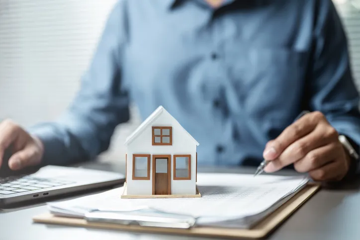Persona firmando documentos junto a una maqueta de casa, representando la gestión y reparto de una herencia inmobiliaria