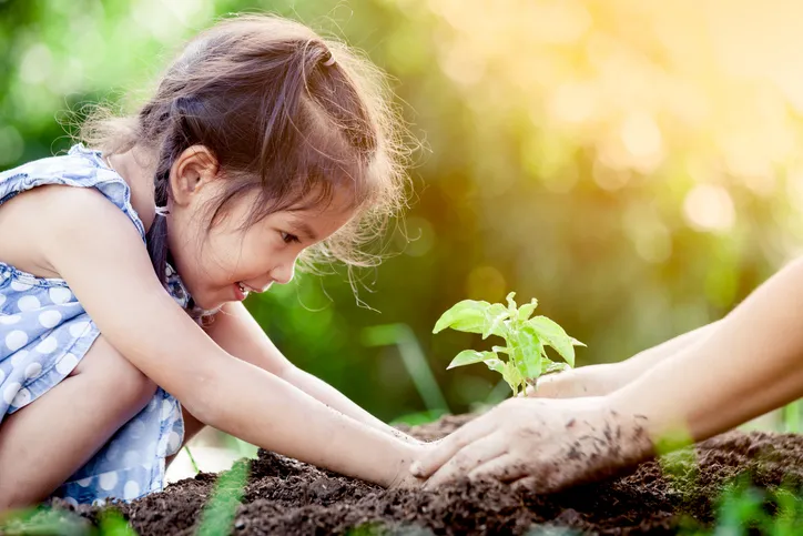 Niña sonriente plantando un brote junto a la mano de un adulto en la tierra, simbolizando el crecimiento o el arraigo familiar.