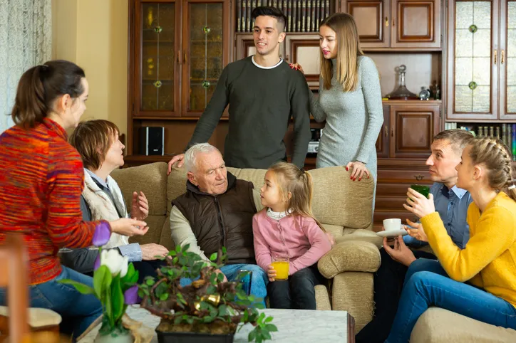 Familia numerosa de varias generaciones reunida y conversando en un salón, incluyendo un abuelo, adultos y una niña pequeña.