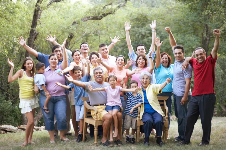 Gran familia numerosa posando y celebrando al aire libre con los brazos levantados, rodeada de árboles.