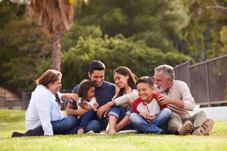 Familia de tres generaciones sentada y riendo sobre el césped de un parque, incluyendo abuelos, padres y dos niños.
