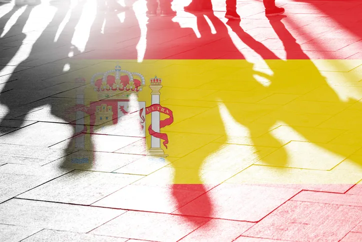 Sombras de personas caminando proyectadas sobre un suelo empedrado, superpuestas con la bandera de España y su escudo.