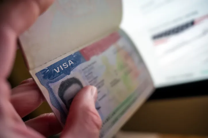 Persona sosteniendo un pasaporte abierto mostrando la página del visado frente a una pantalla de ordenador.
