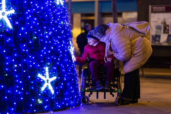 Niño en silla de ruedas sonriendo junto a un adulto frente a un árbol decorado con luces azules navideñas.