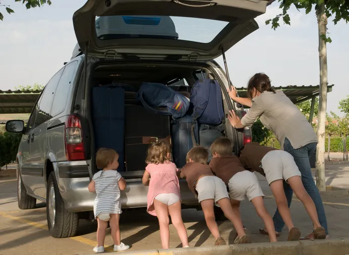 Una madre y varios niños empujando juntos un coche familiar cargado de maletas en un aparcamiento.