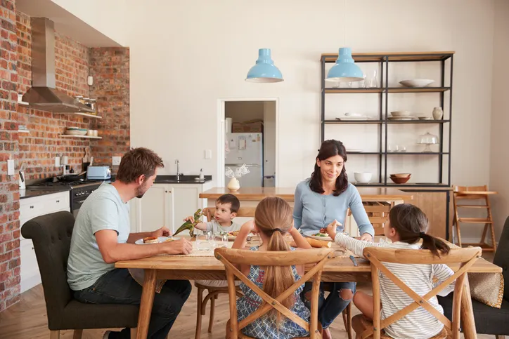 Familia con tres niños sentados a la mesa comiendo juntos en una cocina moderna con pared de ladrillo visto.