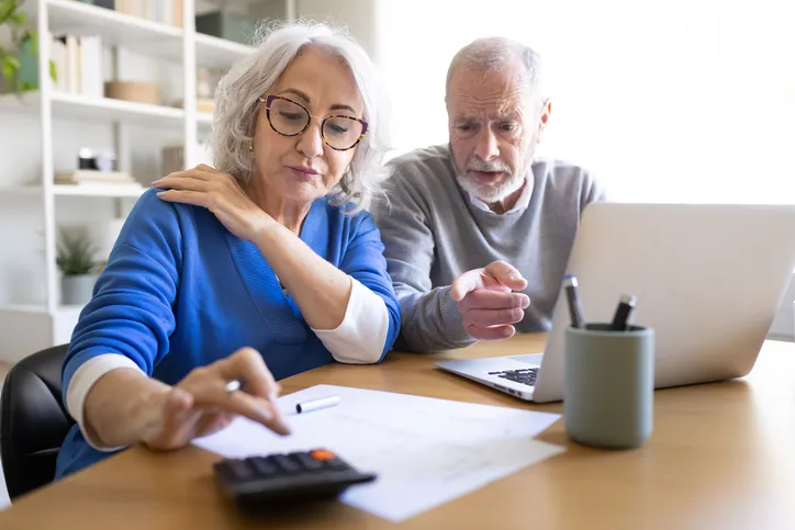 Pareja de jubilados calculando su pensión con calculadora, documentos y ordenador portátil