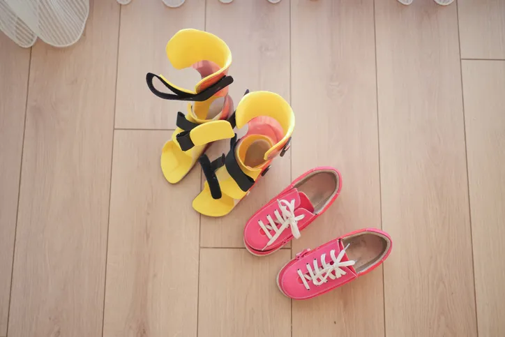 Ortesis ortopédicas infantiles amarillas junto a un par de zapatillas rosas en el suelo