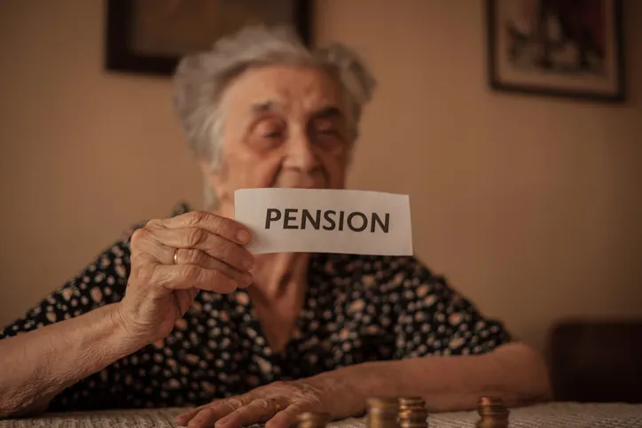 Mujer mayor sosteniendo un cartel con la palabra 'pensión' junto a montones de monedas