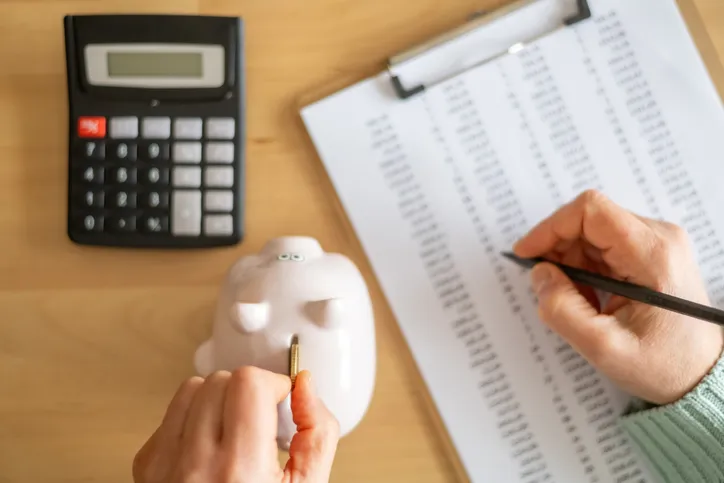 Persona anotando cifras en un listado junto a una calculadora y una hucha con forma de cerdito