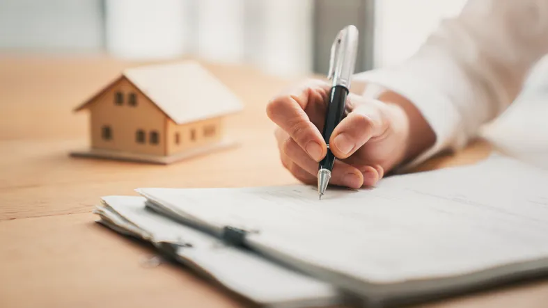 Persona firmando un documento junto a una maqueta de casa, relacionado con la disposición de bienes inmuebles en un testamento