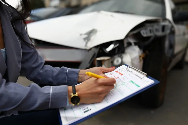 Persona rellenando un informe de siniestro junto a un coche accidentado, tras un choque