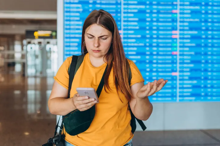Mujer con gesto de frustración mirando el móvil frente al panel de vuelos en un aeropuerto
