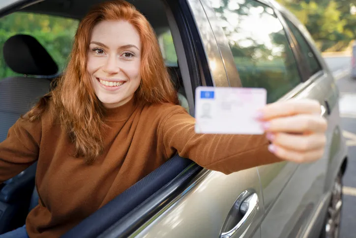 Mujer joven sonriente mostrando su carnet de conducir desde la ventanilla del coche