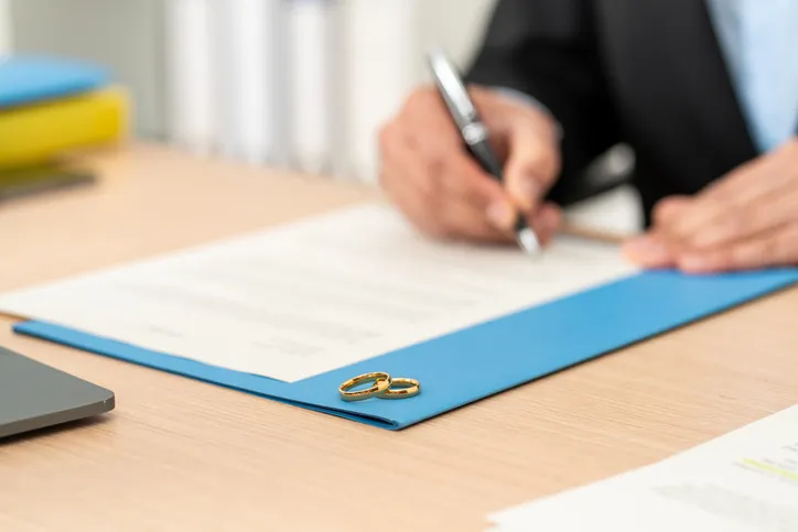 Persona firmando un documento legal sobre una carpeta azul con dos alianzas encima, representando el registro de una pareja de hecho o matrimonio