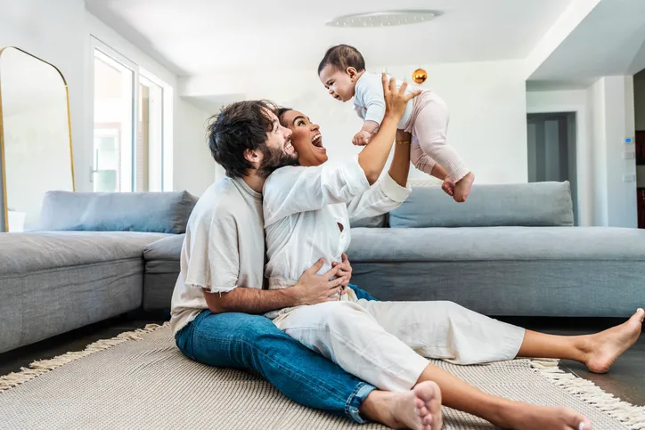 Padres sentados en el suelo del salón sonriendo mientras levantan a un bebé en brazos, reflejando la alegría de formar una familia mediante adopción