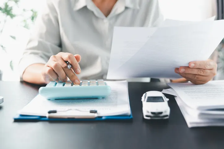 Mujer revisando documentos y usando una calculadora junto a un coche en miniatura, gestionando el seguro del vehículo