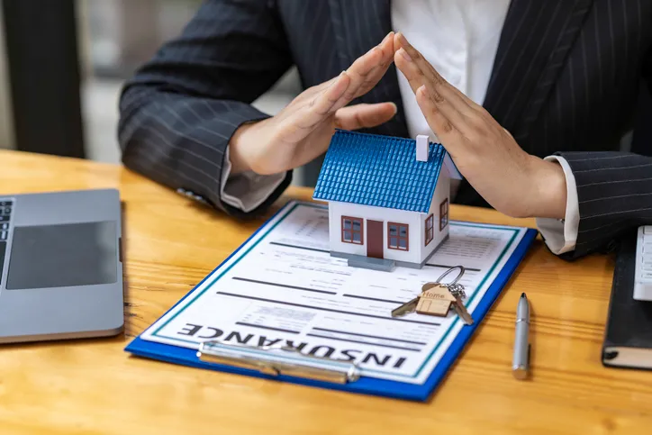 Manos formando un techo protector sobre una casa en miniatura colocada sobre un documento de seguro de hogar