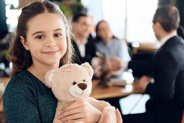 Niña sonriente abrazando un peluche mientras al fondo unos adultos firman documentos en una oficina, relacionado con un proceso de adopción