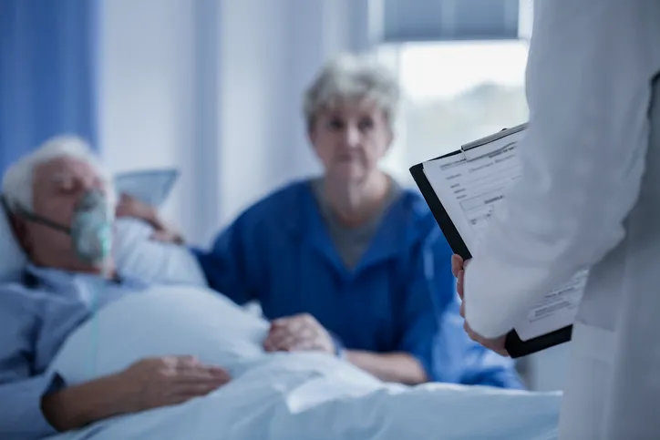 Médico con una tablilla informando a una familiar sobre un paciente hospitalizado con mascarilla de oxígeno
