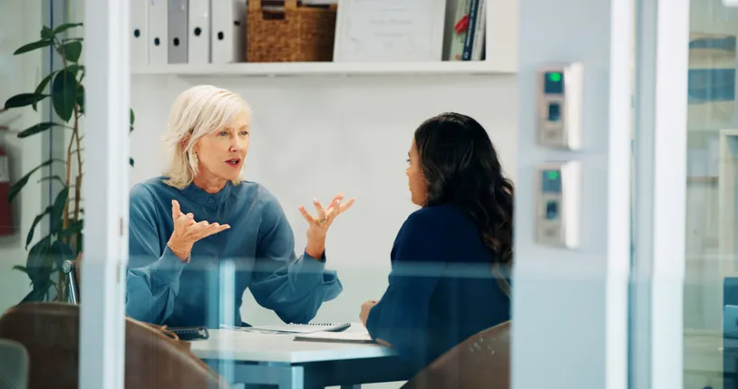 Dos mujeres conversando en una oficina, una explicando algo con las manos a la otra