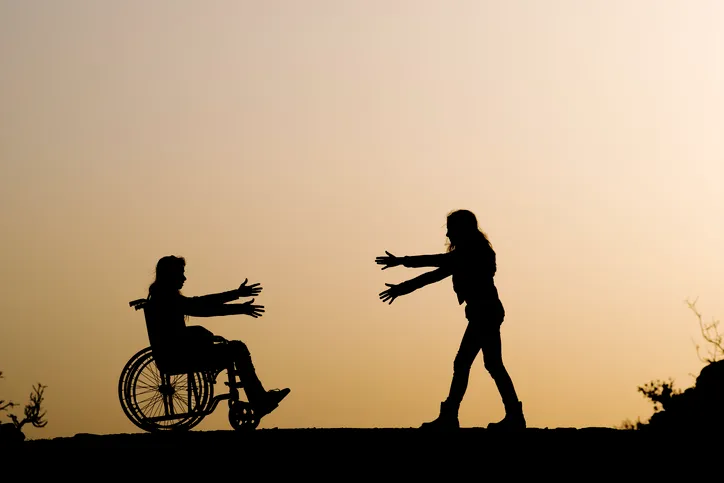 Siluetas al atardecer de una persona en silla de ruedas extendiendo los brazos hacia otra persona que se acerca, representando el apoyo y cuidado a personas con discapacidad