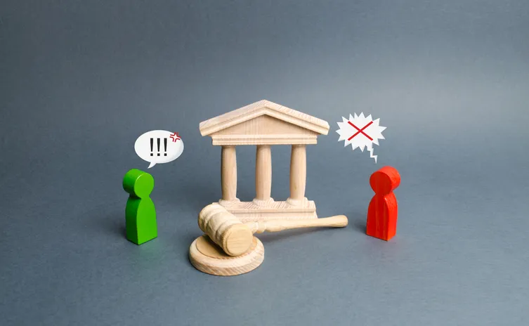 Figuras de madera verde y roja frente a un edificio judicial en miniatura con un mazo de juez, representando un conflicto legal entre denunciante y denunciado.