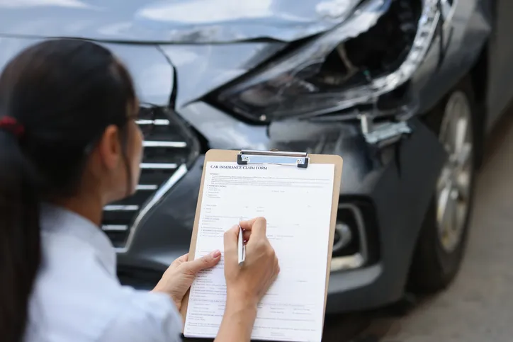 Mujer rellenando un formulario de reclamación de seguro frente a un coche con daños en la parte delantera tras un accidente.