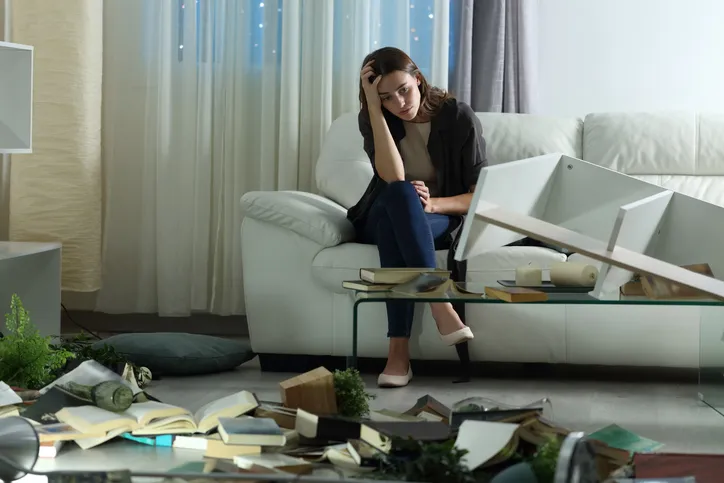Mujer angustiada sentada en un sofá dentro de un salón desordenado, con muebles caídos y libros esparcidos por el suelo tras un robo o allanamiento.