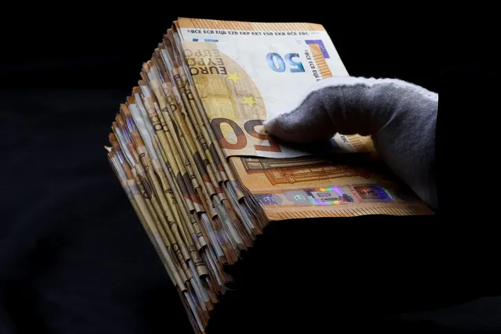 Mano con guante sujetando un fajo de billetes de 50 euros sobre fondo oscuro, sugiriendo dinero de procedencia ilícita o una transacción encubierta.