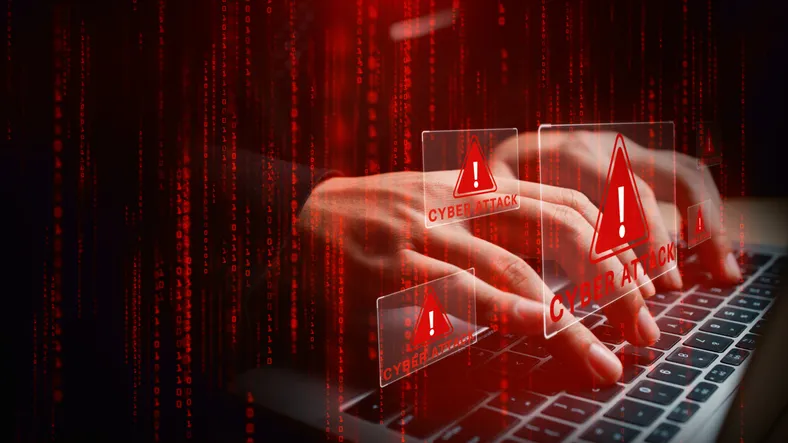 Manos escribiendo en un teclado de portátil con alertas rojas de 'Cyber Attack' y código binario de fondo, representando un ciberataque en curso.